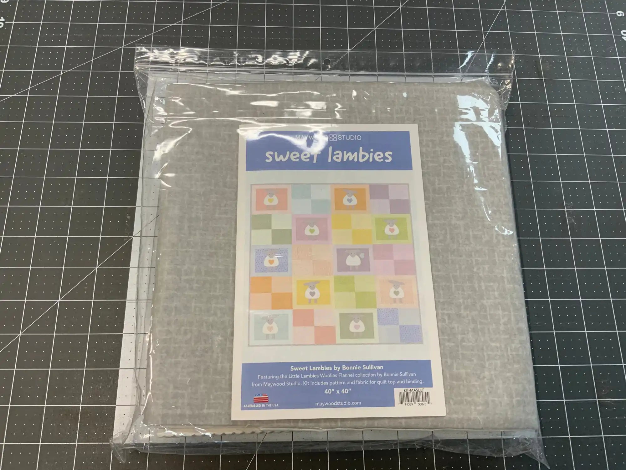 Packaged ’Sweet Lambies’ quilt pattern with gray fabric visible through clear plastic packaging.